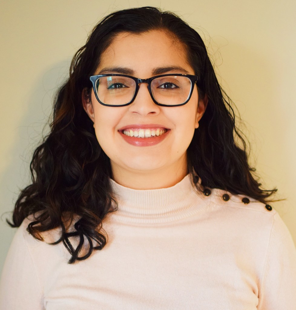 Headshot of Dhruti Bhagat-Conway, a person with light beige skin, from the shoulders up, smiling, in a sweater and glasses, with shoulder-length black wavy hair